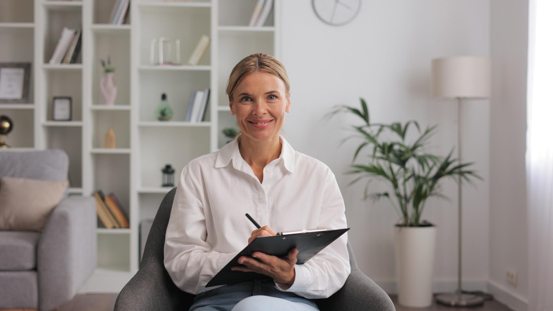 Happy professional psychologist woman sitting in chair writing on clipboard, looking and smiling at camera, working in modern office. Psychotherapeutic services, mental health professional concept.